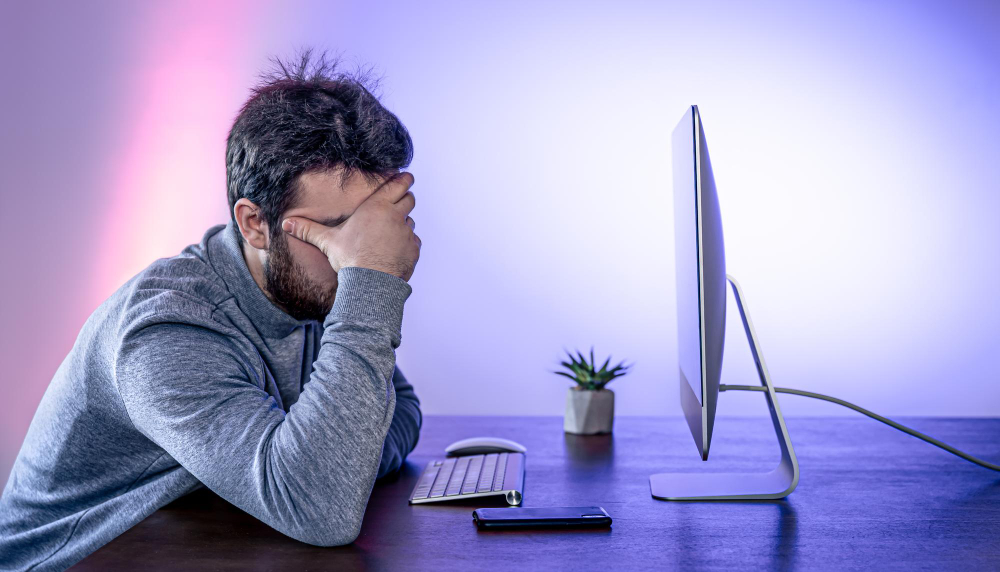 tired man sits front computer covering his face with his hands